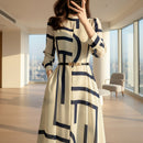 Woman wearing a long white dress with blue geometric patterns, standing in front of a marble wall.