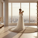 Woman in a white wedding dress holding flowers in a bright room with sheer curtains.