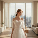Woman wearing a white off-shoulder dress with sheer sleeves in front of white curtains.