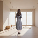 Woman wearing a gray plaid dress standing in a room with beige curtains.