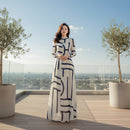 Woman in a long white dress with blue geometric patterns taking a mirror selfie in a modern room.